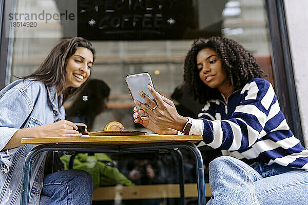 Glückliche junge Frau mit Freundin  die im Straßencafé ihr Smartphone benutzt