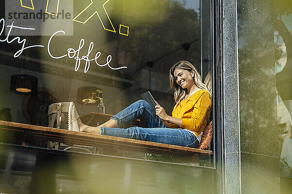 Glückliche Frau mit Tablet-PC sitzt am Fenster im Café