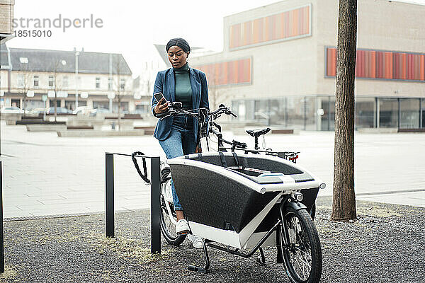 Geschäftsfrau surft mit Smartphone im Internet und sitzt auf einem Lastenfahrrad