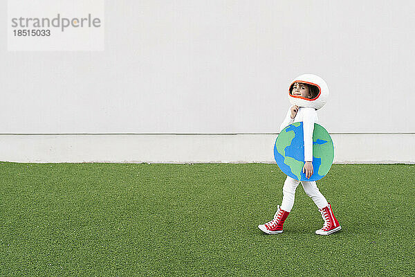 Lächelndes Mädchen im Astronautenkostüm  das die Erde in der Hand hält und auf Gras läuft