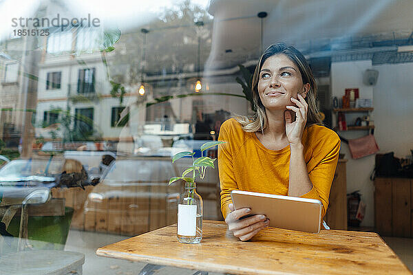 Lächelnde Frau mit Tablet-PC sitzt am Tisch im Café und sieht durch Glas