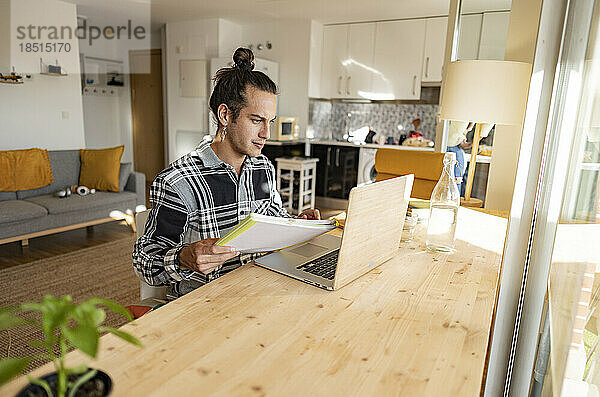 Junger Mann mit Dokument  der im Heimbüro am Laptop arbeitet