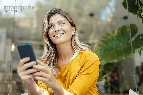 Glückliche Frau mit blonden Haaren hält Smartphone vor Glaswand