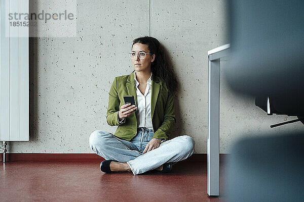 Nachdenkliche junge Geschäftsfrau sitzt mit Mobiltelefon im Büro