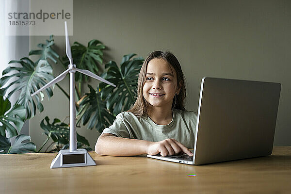 Lächelndes Mädchen mit Laptop und Windturbinenmodell auf dem heimischen Tisch