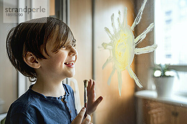 Glücklicher Junge mit Pinsel  der Sonne auf Glas zeichnet
