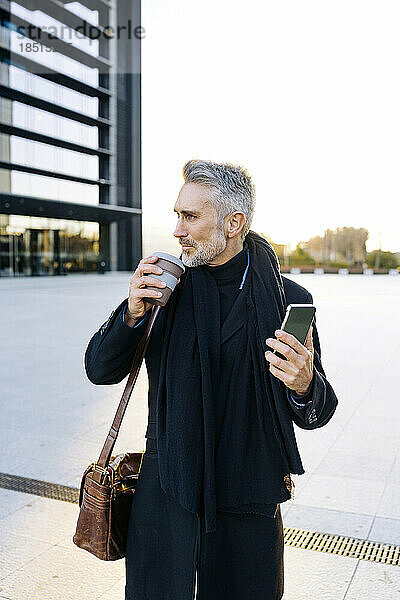 Reifer Geschäftsmann steht mit Kaffeetasse und Smartphone am Fußweg