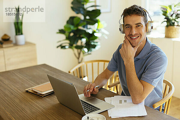 Glücklicher Freiberufler mit Kopfhörern  der mit Laptop am Tisch im Heimbüro sitzt