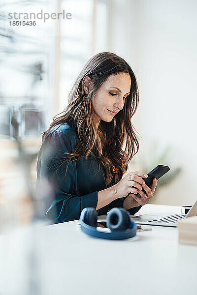 Lächelnde junge Geschäftsfrau  die im Heimbüro ihr Smartphone benutzt