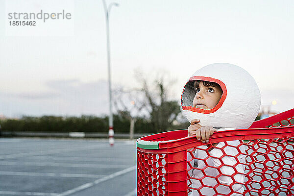Nachdenkliches Mädchen im Astronautenkostüm sitzt im Einkaufswagen