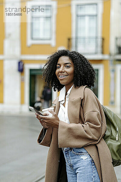 Glückliche junge Frau mit Smartphone und Kaffeetasse steht vor dem Gebäude