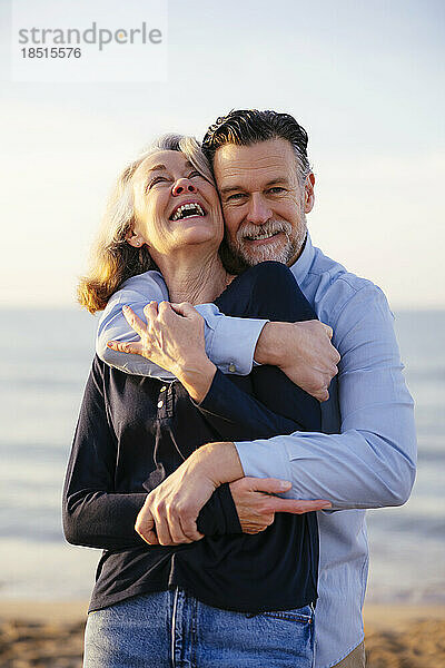 Glücklicher Mann umarmt fröhliche reife Frau am Strand
