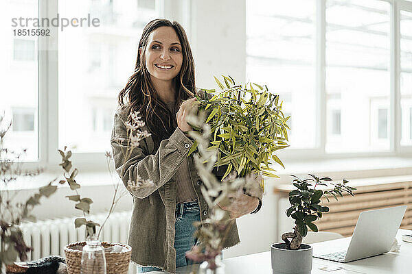 Glückliche Geschäftsfrau steht mit Topfpflanze im Heimbüro