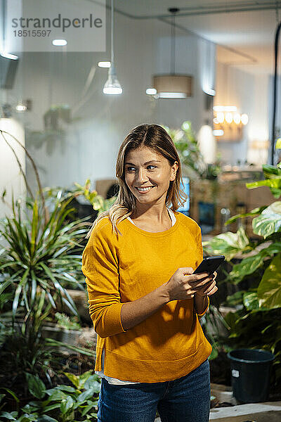 Lächelnde Geschäftsfrau mit Smartphone steht vor Pflanzen im Büro