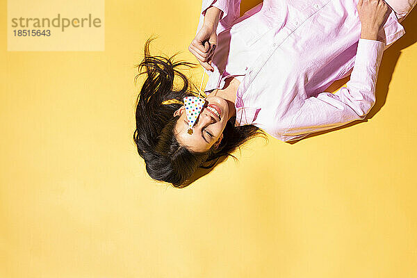 Happy woman covering eye with prop lying against yellow background