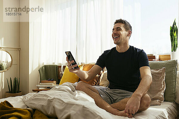 Lachender Mann benutzt Smartphone und sitzt mit gekreuzten Beinen zu Hause im Bett
