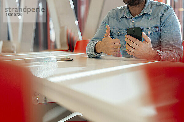 Geschäftsmann mit Smartphone zeigt Daumen nach oben und sitzt am Tisch