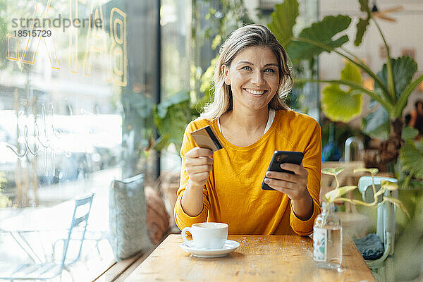 Glückliche Frau sitzt mit Kreditkarte und Smartphone im Café