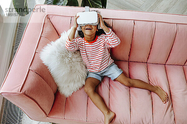 Happy girl wearing virtual reality simulator sitting on sofa