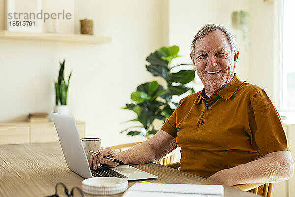 Glücklicher älterer Mann  der zu Hause mit Laptop am Tisch sitzt