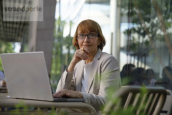 Nachdenkliche Geschäftsfrau mit der Hand am Kinn sitzt mit Laptop im Straßencafé