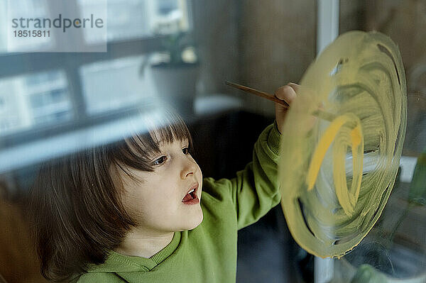 Cute boy drawing sun on glass with paintbrush at home