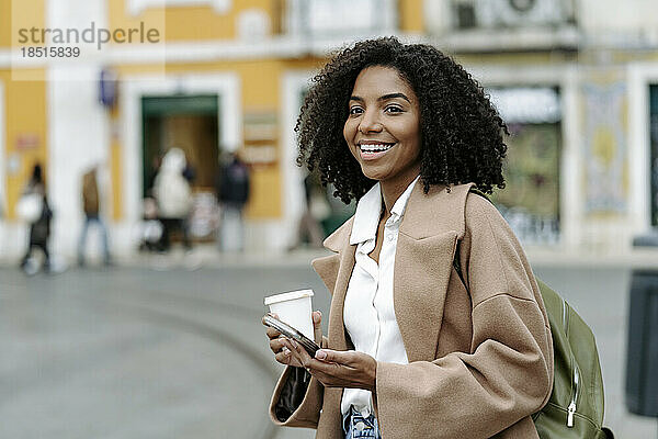 Glückliche junge Frau steht mit Kaffeetasse und Mobiltelefon