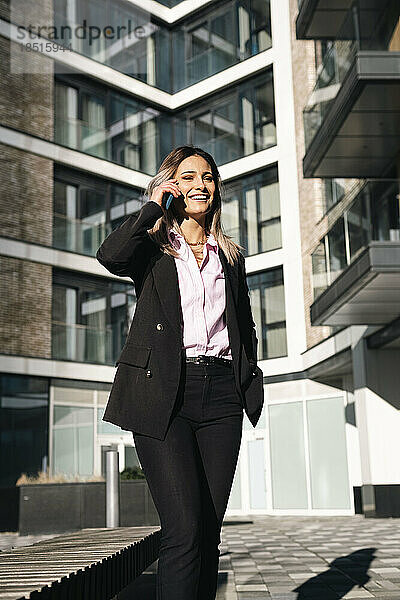 Lächelnde Geschäftsfrau  die vor dem Bürogebäude mit dem Smartphone spricht