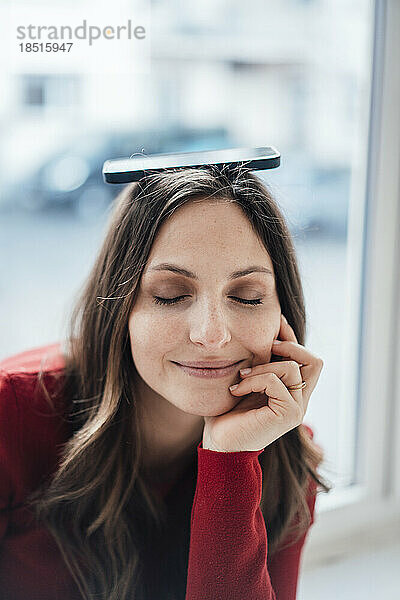 Frau mit geschlossenen Augen balanciert ihr Smartphone auf dem Kopf vor dem Fenster