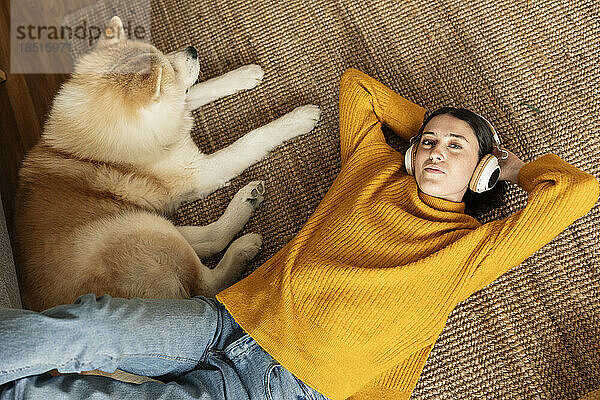 Junge Frau mit kabellosen Kopfhörern hört Musik und liegt neben Hund auf dem Teppich