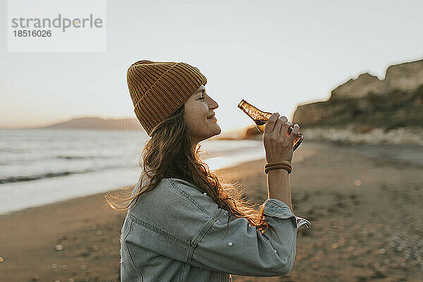 Lächelnde Frau mit Bierflasche steht am Strand