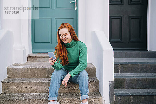 Glückliche junge Frau  die ihr Smartphone benutzt und auf einer Treppe vor dem Haus sitzt