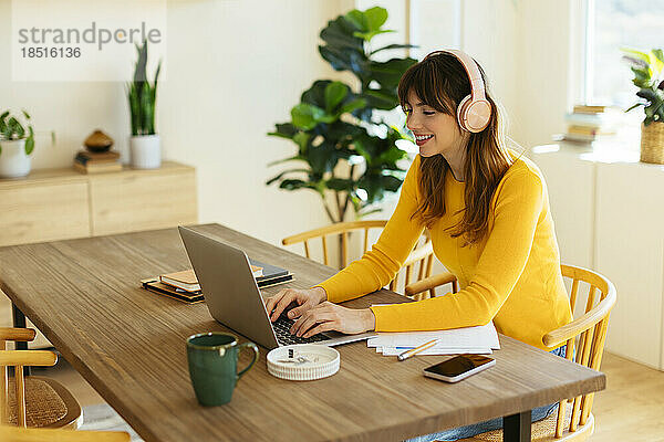 Glückliche Frau mit Kopfhörern  die im Heimbüro am Laptop arbeitet