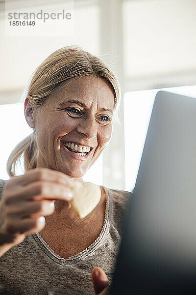 Glücklicher Freiberufler  der im Heimbüro einen Snack isst und am Laptop arbeitet