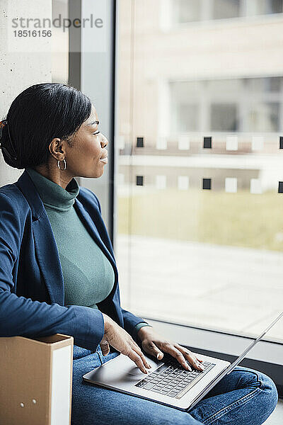Geschäftsfrau schaut aus dem Fenster und sitzt mit Laptop im Büro