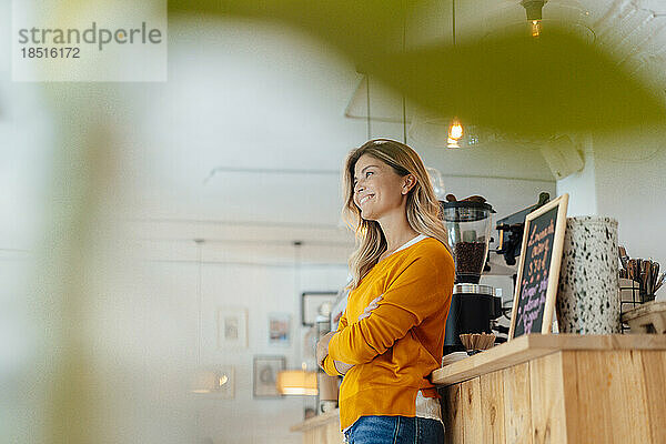Thoughtful smiling woman standing with arms crossed in cafe