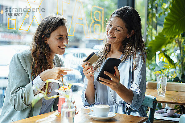 Glückliche Frau mit Kreditkarte zeigt einem Freund im Café ihr Smartphone