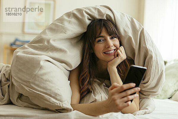 Glückliche Frau mit Handy zu Hause in Decke gehüllt
