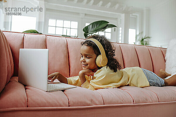 Lächelndes Mädchen  das auf dem Sofa liegt und zu Hause im Wohnzimmer am Laptop lernt