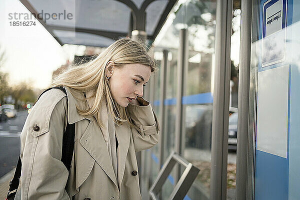 Blonde junge Frau überprüft den Busfahrplan am Bahnhof