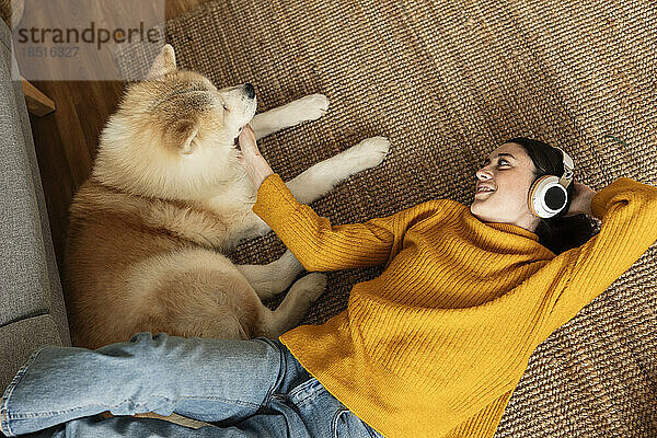Smiling woman wearing wireless headphones listening to music and stroking dog on carpet at home