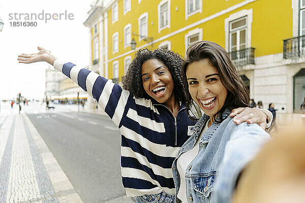 Fröhliche Frau macht ein Selfie mit einer Freundin auf der Straße vor einem gelben Gebäude