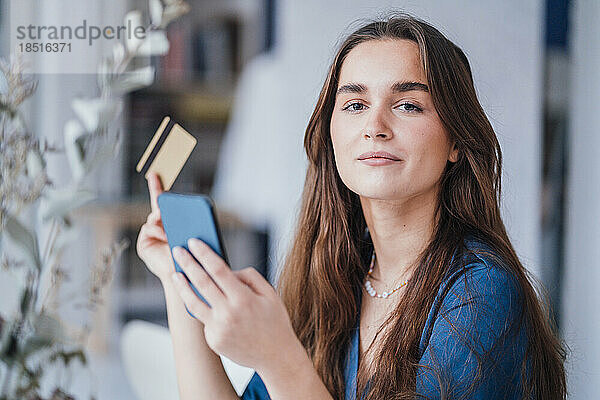 Confident young businesswoman with mobile phone and credit card in office