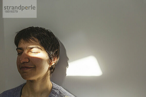 Lächelnde Frau genießt Sonnenlicht auf ihrem Gesicht vor der Wand