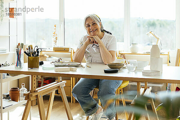 Glückliche ältere Frau sitzt mit der Hand am Kinn am Tisch in der Werkstatt