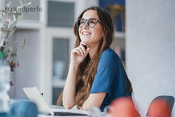 Lächelnde junge Geschäftsfrau mit der Hand am Kinn sitzt am Schreibtisch