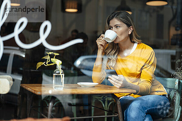 Frau mit Smartphone trinkt Kaffee im Café  gesehen durch Glas