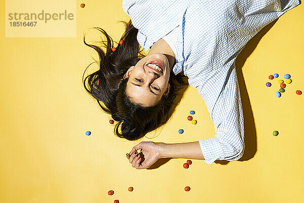 Happy young woman lying amidst colorful candies against colored background