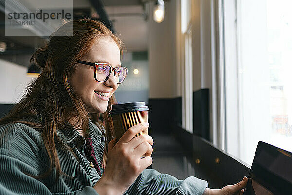 Fröhlicher junger Freiberufler mit Einweg-Kaffeetasse sitzt im Café