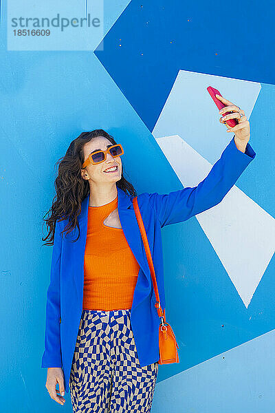 Glückliche Frau mit Sonnenbrille macht ein Selfie vor einer blauen Wand